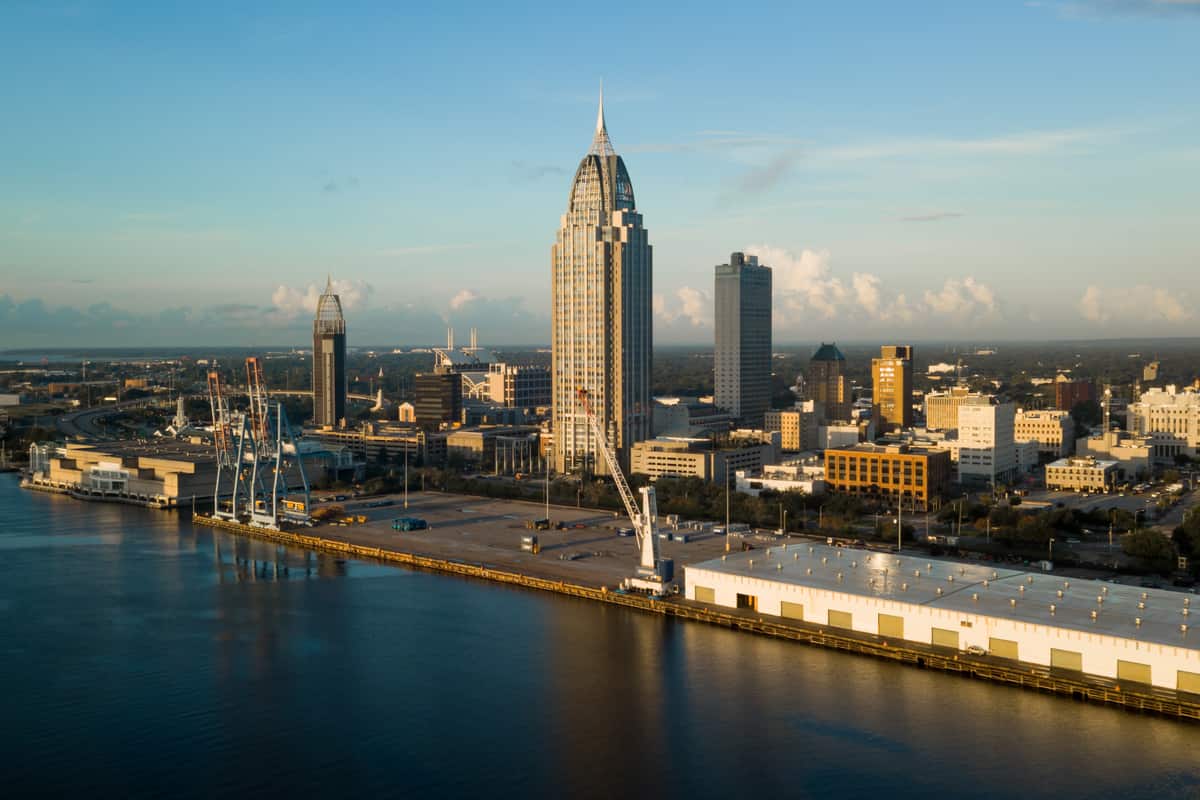 The spire of the RSA Battle House Tower rising over downtown Mobile, Alabama, with port gantry cranes and a working wharf along the Mobile River in the foreground